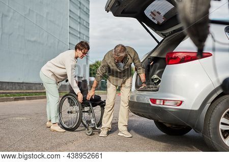Full Length Portrait Of Mature Couple Unloading Wheelchair Out Of Car Trunk In Parking Lot Outdoors,
