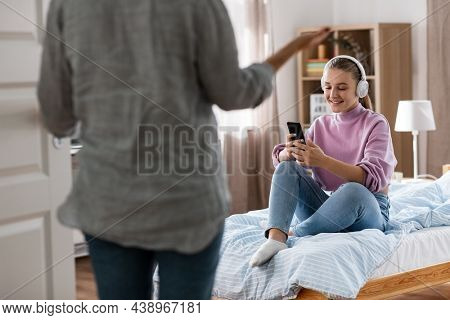 misbehavior, conflict and family concept - angry mother entering room and smiling daughter in headphones with smartphone sitting on bed at home