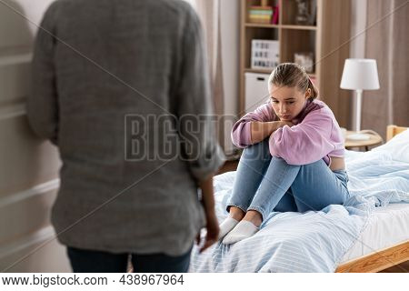 misbehavior, conflict and family concept - mother entering room and sad daughter sitting on bed at home