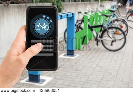 technology, transport and sustainability concept - close up of hand holding smartphone with mobile app and bicycle icon on screen over electric bike parking and charging station in city on background