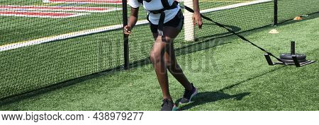 A High School Girl Track Runner Is Pulling A Sled With Weights On A Turf Field Next To A Protective 