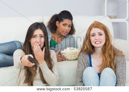 Portrait of scared young female friends with remote control and popcorn bowl on sofa at home