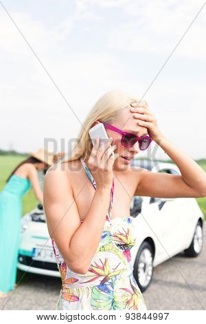 Frustrated woman using cell phone while friend examining broken down car at countryside