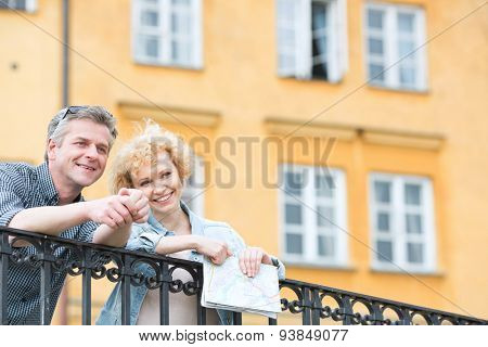 Happy middle-aged couple with map leaning on railing against building