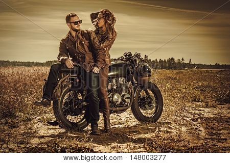 Young, stylish cafe racer couple on the vintage custom motorcycles in a field.