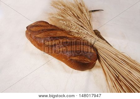 bunch of mown wheat ears with vintage handmade reaper hook sickle and rye dark white french fresh bread loaf on light linen tablecloth