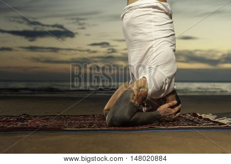 Cropped shot of Young tattoo man doing advanced yoga exercise on the beach at sunset. Practicing head or shoulders stand outdoors.