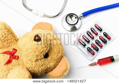 Doctor work table white background desk with medical blister of red and black capsule red injection syringe stethoscope and brown teddy bear. Children medical concept.