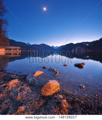 Alpsee lake in Germany at night in spring. Beautiful night landscape with lake mountains forest stars full moon sky and stones in water. Panoramic photo
