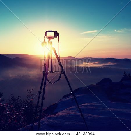 Tripod with running camera on peak ready for photography. Sharp autumn rocky peaks increased from gold fog. Vivid and strong vignetting effect.