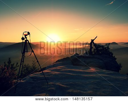 Tripod with running camera on peak ready for photography. Sharp autumn rocky peaks increased from gold fog. Vivid and strong vignetting effect.