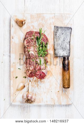 Raw uncooked roastbeef meat cut with rosemary, thyme and garlic and butcher knife on old white painted wooden background, top view, vertical composition