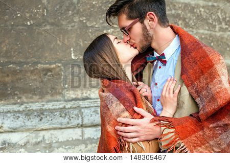 Young fashion elegant stylish couple standing, embracing, wrarps in pled and kiss smb's lips on streets of old european city