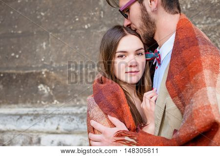 Young fashion elegant stylish couple standing, embracing, wrarps in pled on streets of old european city