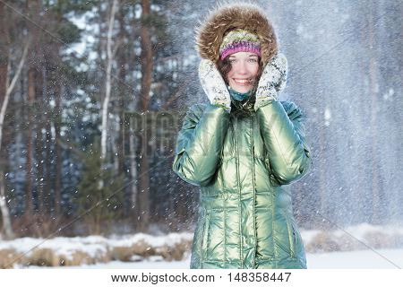 Laughing young woman is holding hands in woolly mittens near face during snowstorm in winter forest outdoors