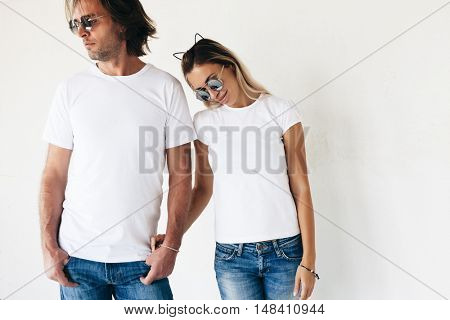 Two hipster models man and woman wearing blanc t-shirt, jeans and sunglasses posing against white wall, toned photo, front tshirt mockup for couple