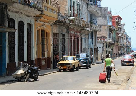 CUBA HAVANA - MAY 14 2012: Old Havana street with vintage American cars. A tourist with a suitcase walking down the street toward the Malecon.