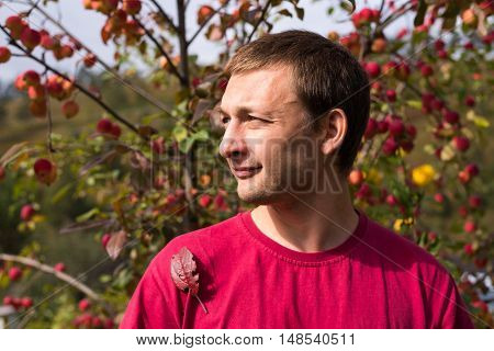 Young man in autumn. Portrait of a young handsome man in a red T-shirt on a sunny day against an apple rennet tree. The sunshine is strong, the shadows are deep.