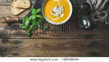Pumpkin cream soup in bowl with fresh basil, spices and grilled bread slices over old rustic wooden background, top view, copy space, horizontal composition