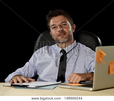 corporate portrait of happy successful businessman in shirt and tie smiling at office desk working with laptop computer isolated on black background confident and trustful in business success concept