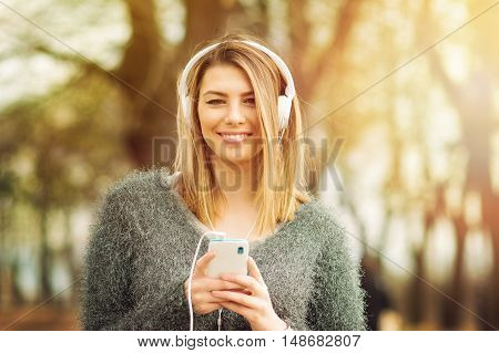 Beautiful young blonde millennial teenage girl with headphones and smart phone in park in autumn. Cool young woman listening to music outdoors in fall.