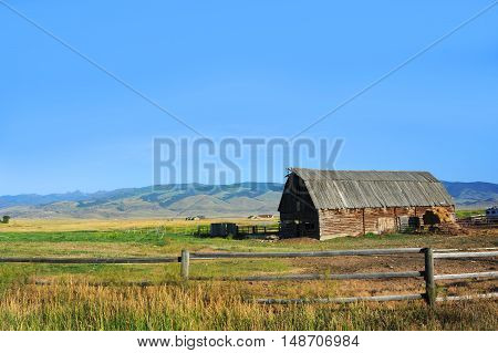 Rustic pole barn sits in a field in Happy Valley Montana. Wooden fence sets off coral. Roof of barn is wooden.