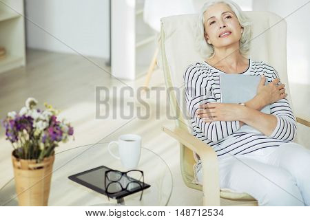 Find peace in mind. Delighted pleasant aged woman crossing her arms and sitting in the arm chair while resting at home