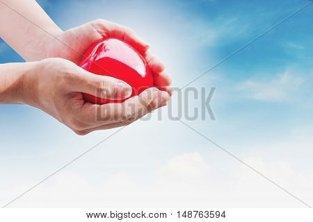 Hand holding red hart on blue sky and white clouds, with bright light