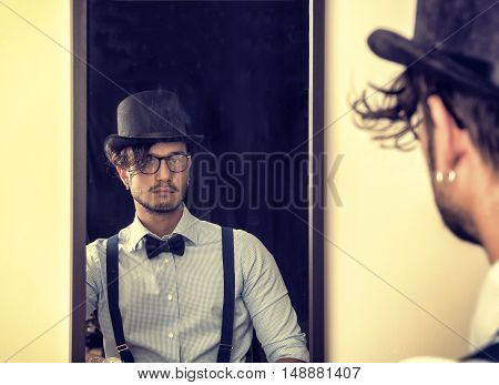 Portrait of brunette young man in glasses, hat, bow-tie, suspenders and shirt looking at himself in a mirror. Vintage look