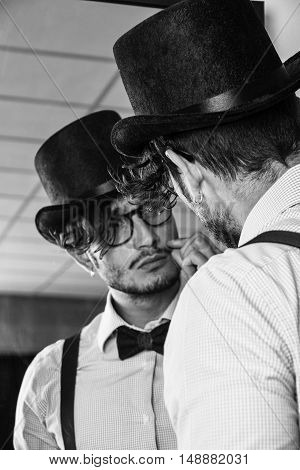 Portrait of brunette young man in glasses, hat, bow-tie, suspenders and shirt looking at himself in a mirror. Vintage look