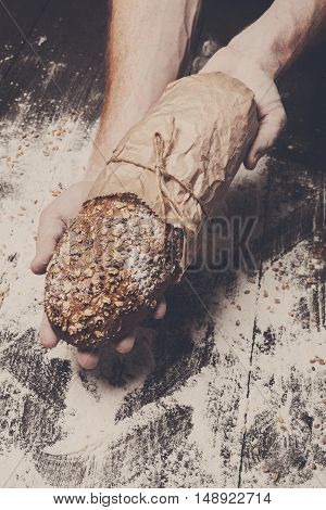 Warm fresh rye whole grain bread, wrapped in craft paper. Healthy baking and cooking concept background. Hands carefully hold loaf on wooden table background, sprinkled with flour. Soft toning
