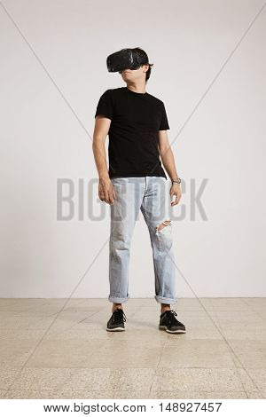 Full body shot of a young male model in VR headset, black unlabeled t-shirt and blue torn jeans looking around the room with white walls and light wooden floor