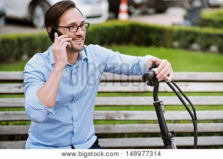 Joyful conversation. Handsome bespectacled man speaking on cellphone and smiling while sitting on the bench.