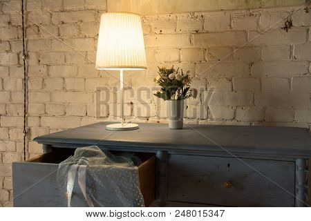 Lamp And Bouquet With Flowers On An Old Chest Of Drawers