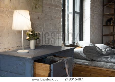 Lamp With A Wooden Chest Of Drawers For Linen And A Bed With Cushions By The Window