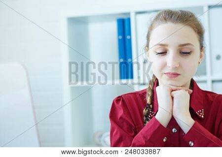 Pensive Young Business Woman Sitting At The Dask With Desktop Monitor And Looking Down At The Papers