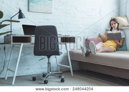 Teen Girl With Cup Of Coffee Reading Book On Fosa In Room With Table And Laptop