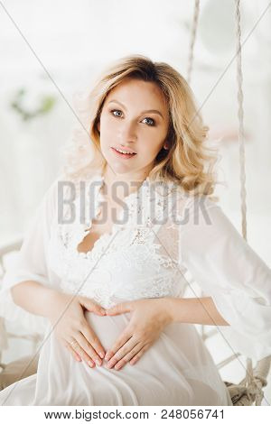 Beautiful Blonde Pregnant Woman Wearing In White Long Dress Sitting At Swing With Flowers And Lookin