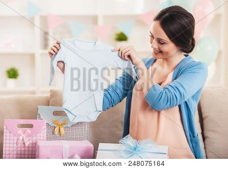 Smiling Pregnant Woman With Blue Baby Romper. Happy Pregnant Mother Holding Romper In Bedroom At Hom