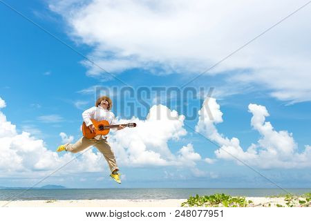 Asian Man Enjoying Summer, Playing Classic Guitar And Jumping On The Beach In Vacations, Blue Sky Ba