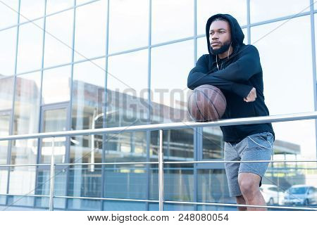 Pensive African American Man In Earphones Leaning At Railings With Basketball Ball And Looking Away