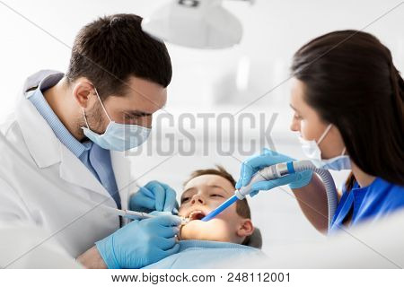 medicine, dentistry and healthcare concept - dentist and assistant with dental drill and saliva ejector treating kid patient teeth at dental clinic