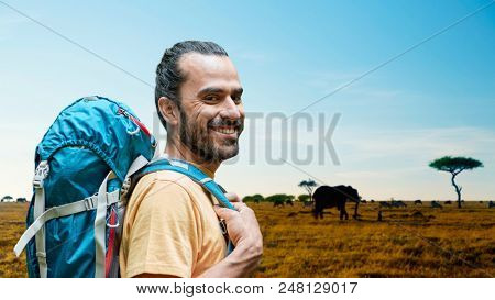 adventure, travel, tourism, hike and people concept - smiling man with backpack over anilmals in african savannah background