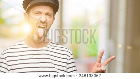 Sailor captain man smoking a tobacco pipe looking at camera showing tong and making victory sign with fingers