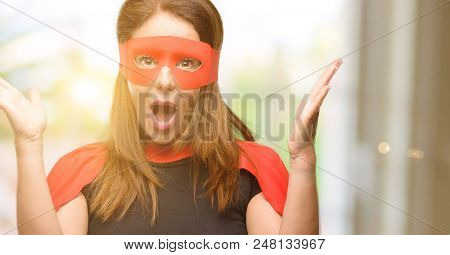 Middle age super hero woman wearing red mask and cape happy and surprised cheering expressing wow gesture