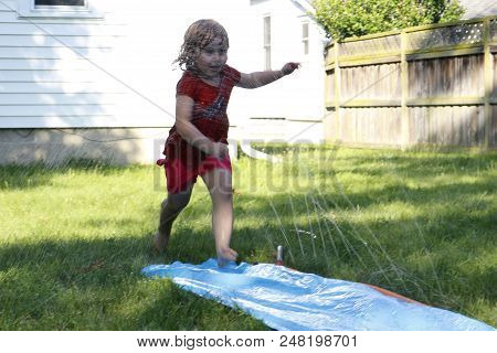Little Preschool Girl Runs Through A Backyard Slip And Slide Water Slide. A Fun Backyard Activity Fo