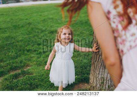 Smiling Little Cute Child Baby Girl In Light Dress Lean On Tree, Looking On Mother On Green Grass In