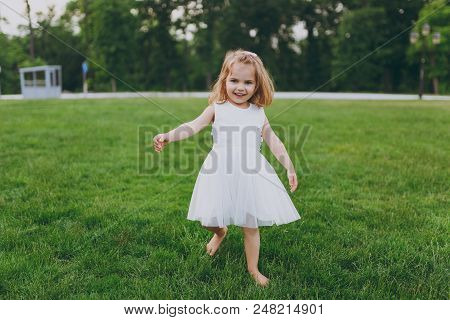 Smiling Little Cute Child Baby Girl In Light Dress Walking And Running, Have Fun On Green Grass Lawn