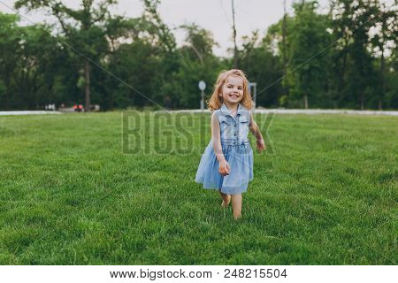 Smiling Little Cute Child Baby Girl In Denim Dress Walking And Running, Have Fun On Green Grass Lawn