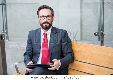 Relaxed Businessman In Glasses Using His Pc Tablet While Sitting On Bench. Senior Businessman Using 